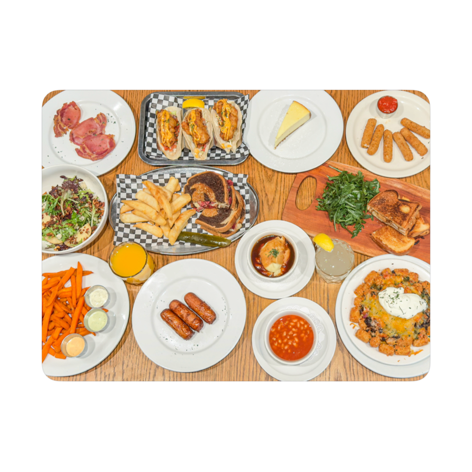 Overhead view of a wooden table covered with assorted brunch dishes, including eggs, sausages, fries, toast, salads, and drinks.