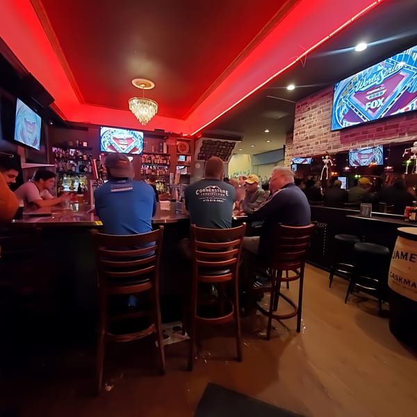 Patrons seated at a bar with red lighting, multiple TVs, and a Jameson whiskey barrel.