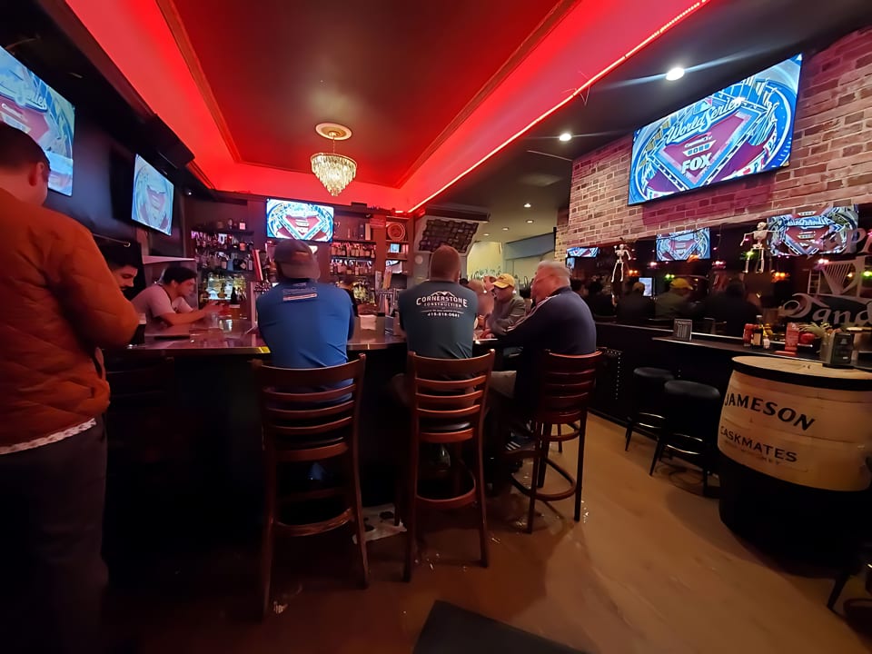Patrons seated at a bar with red lighting, multiple TVs, and a Jameson whiskey barrel.