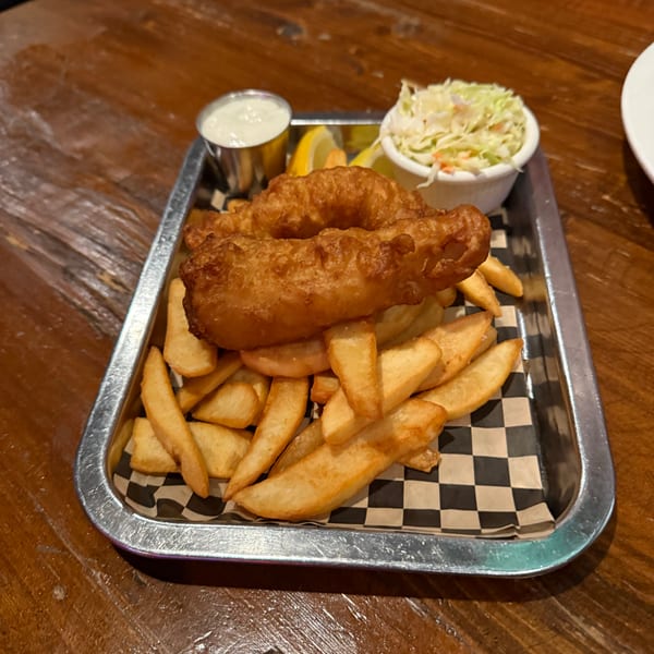 Golden fried fish and chips with coleslaw, tartar sauce, and lemon wedges on a metal tray.