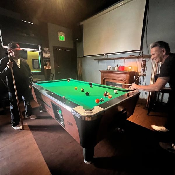 Two men playing pool in a dimly lit bar with a projector screen and fireplace.