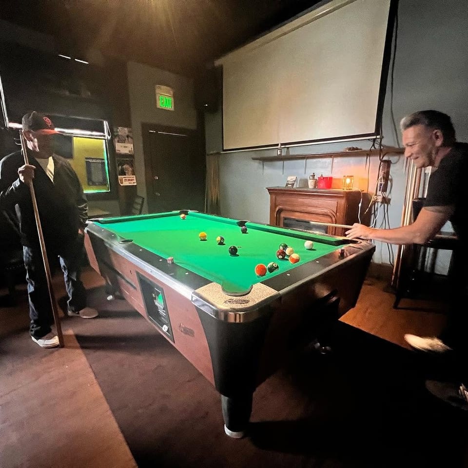 Two men playing pool in a dimly lit bar with a projector screen and fireplace.