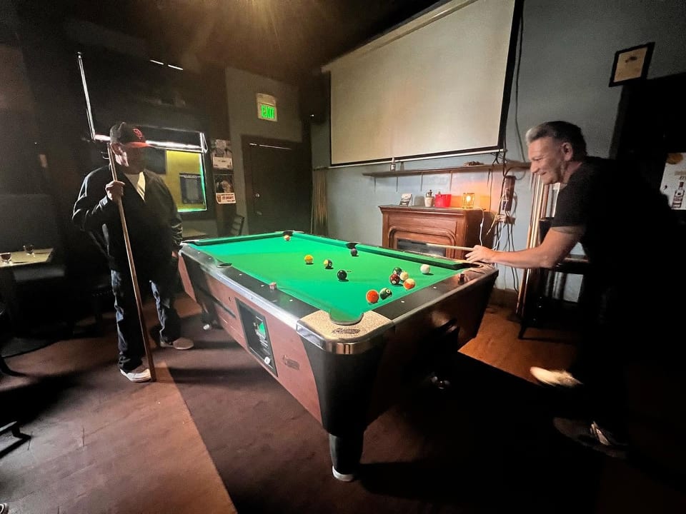 Two men playing pool in a dimly lit bar with a projector screen and fireplace.
