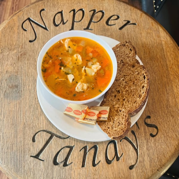 A bowl of chicken and vegetable soup with two slices of brown bread and butter on a wooden table.