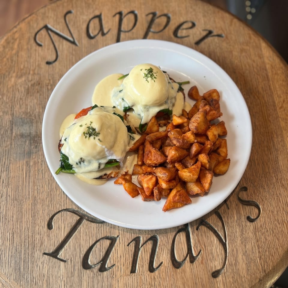 Eggs Benedict with hollandaise sauce and a side of roasted potatoes on a white plate.