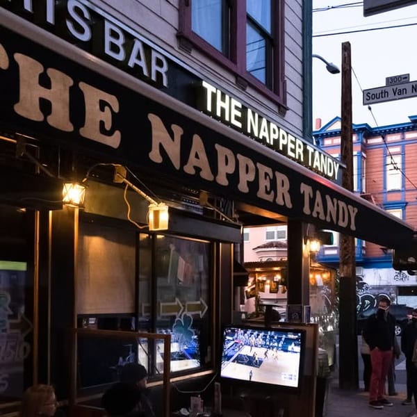 Exterior of The Napper Tandy bar at dusk, with people on the sidewalk and a TV showing sports.