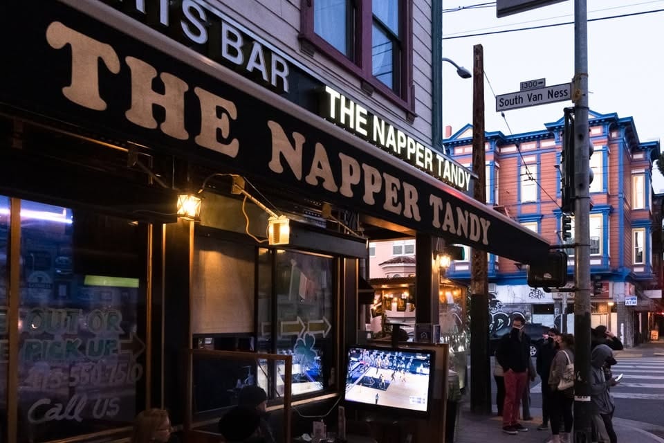 Exterior of The Napper Tandy bar at dusk, with people on the sidewalk and a TV showing sports.