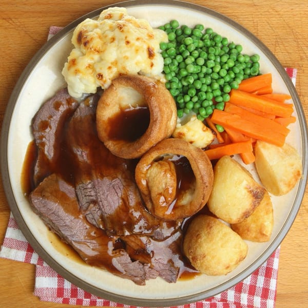 A plate of roast beef with gravy, Yorkshire puddings, roasted potatoes, peas, carrots, and cauliflower cheese.