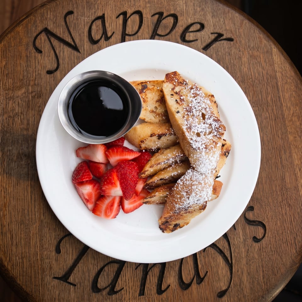 French toast sticks with powdered sugar, a side of strawberries, and syrup on a white plate.